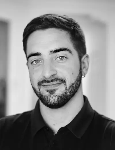 Hombre sonriente con cabello oscuro y barba, camisa polo negra.